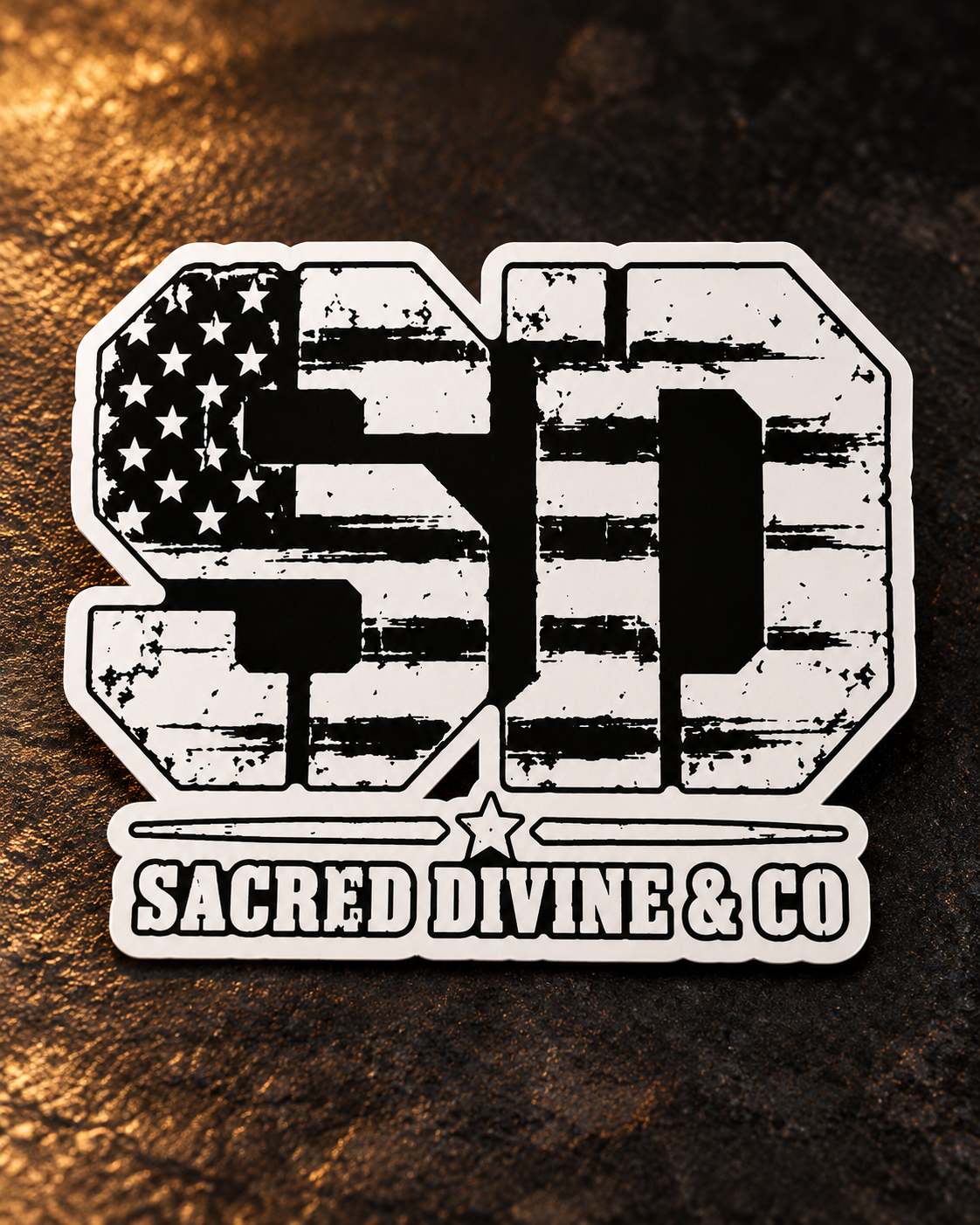 Distressed American Flag SD vinyl decal with Sacred Divine and Co lettering and a white border on a dark textured surface.