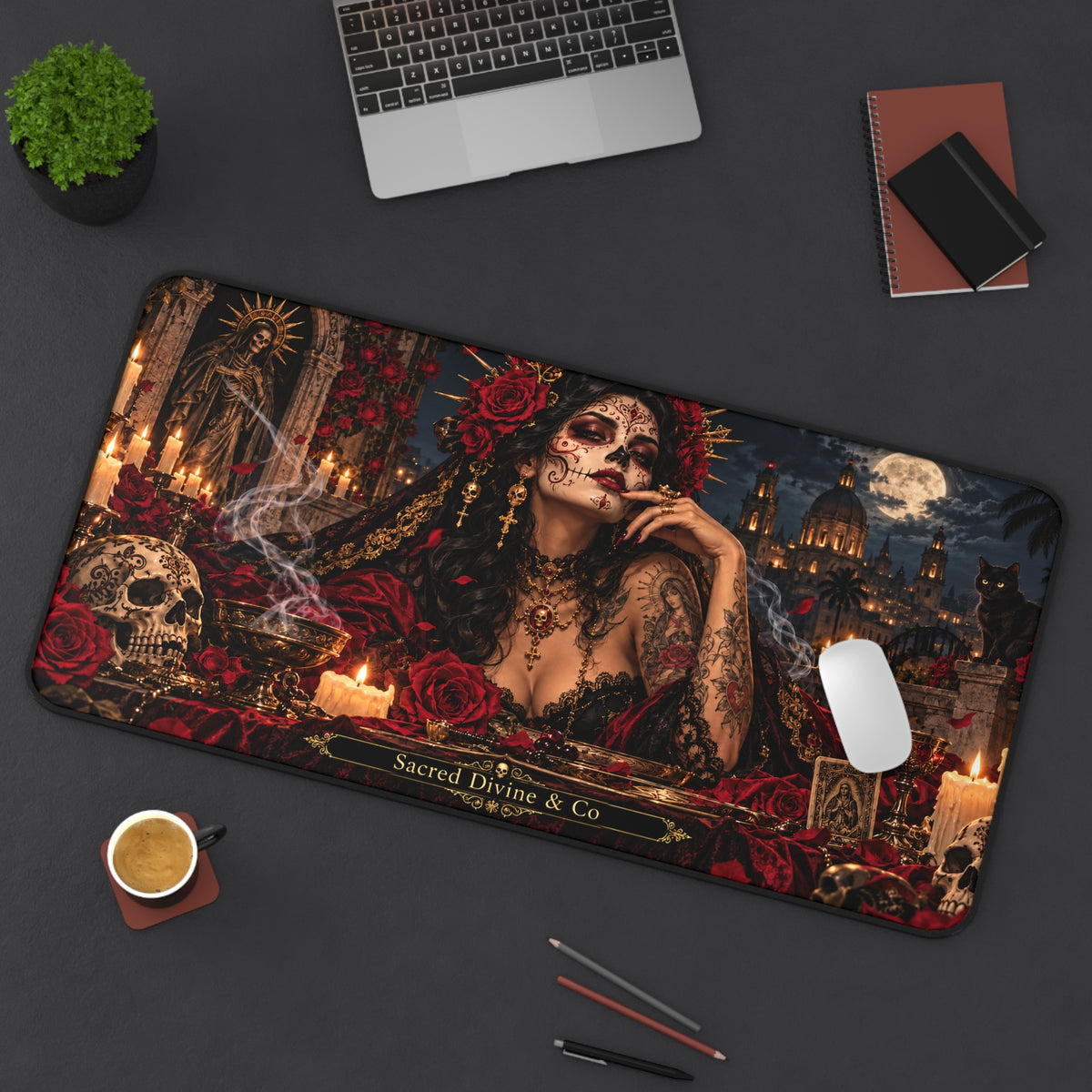 Angled view of the Gothic Day of the Dead desk mat showing sugar-skull woman with red roses against moonlit architecture, with a laptop, plant, notebook, coffee, and pencils on the desk.
