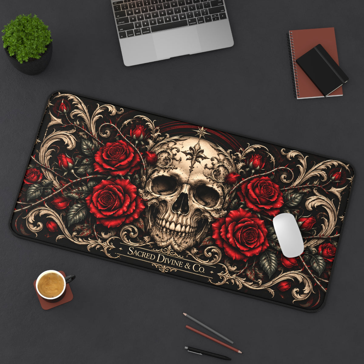 Angled view of the Gothic Skull and Red Roses desk mat showing centered skull framed by ornate cream scrollwork, with a laptop, plant, notebook, coffee, and pencils on the desk.