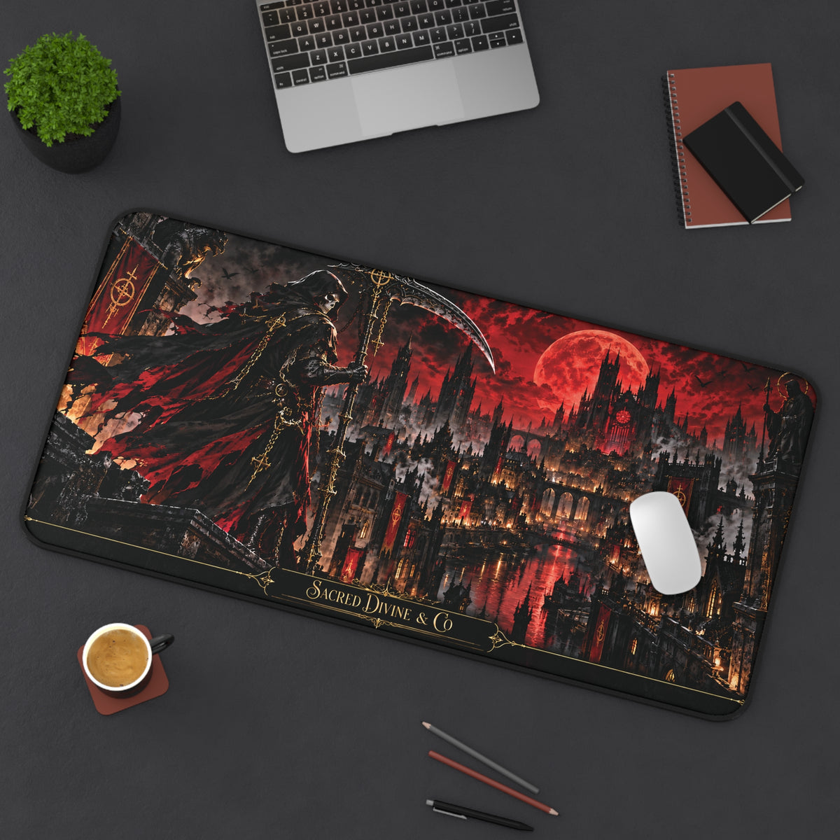 Angled view of the Gothic Reaper Castle desk mat showing grim reaper and dark castle beneath a crimson moon, with a laptop, plant, notebook, coffee, and pencils on the desk.
